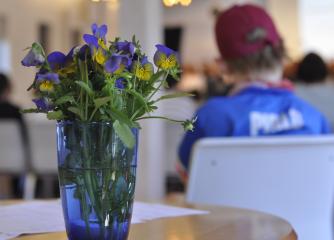 En bukett med lila och gula blommor i en vas och i bakgrunden en person med blå tröja