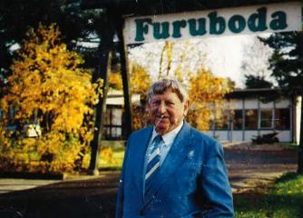 En äldre bild på en man i blå kostym framför en Furuboda-skylt
